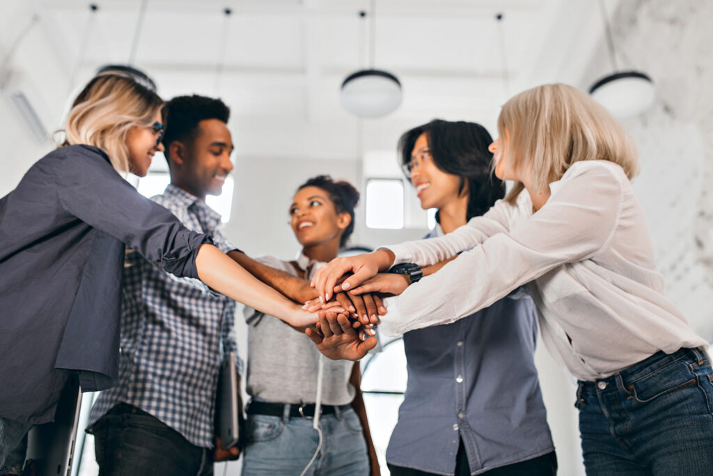 Cheerful international students with happy face expression going to work together on science project. Indoor photo of blonde woman in trendy blouse holding hands with coworkers..